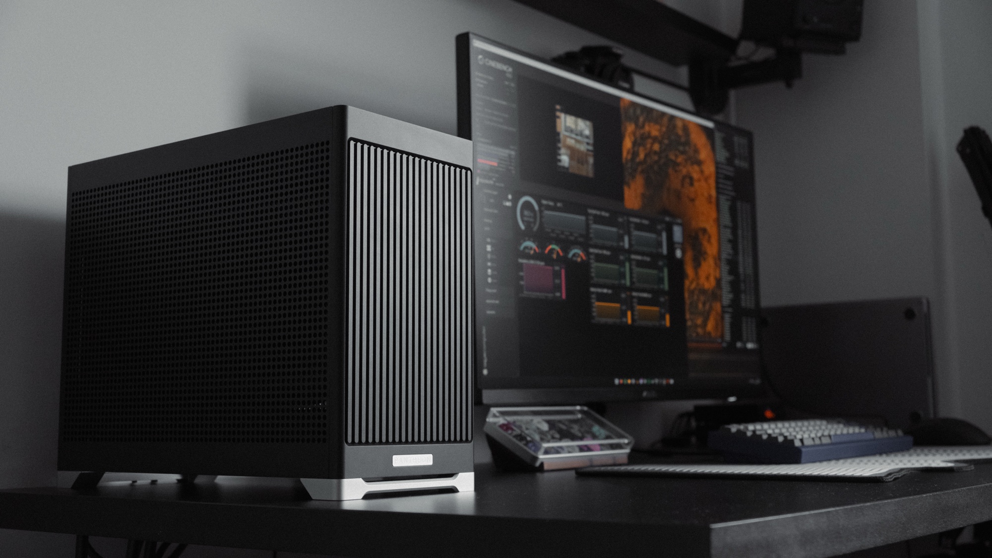 The Pantheon on a desk beside a monitor, mid benchmark